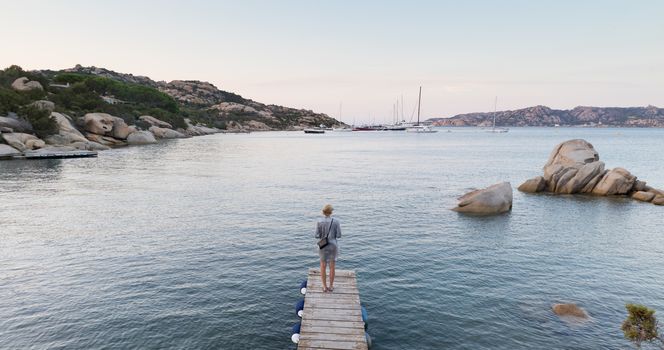 Beautiful woman in luxury summer dress standing on wooden pier enjoying peaceful seascape at dusk. Female traveler stands on a wooden pier in Porto Rafael, Costa Smeralda, Sardinia, Italy.