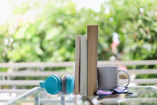Education and relaxation Concept. Closeup of white  mug cup of hot coffee, light blue headphone, reading glasses and books on glass table in green garden view