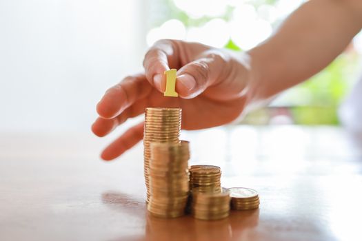 Business, Money, Finance, Secure and Saving Concept. Close up of woman hand holding yellow wooden number and put to top of stack of coins on wooden table with copy space.