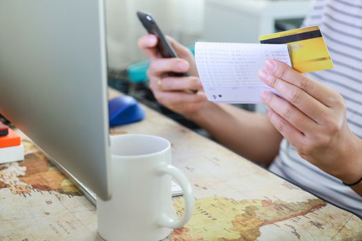 E-Commerce, Shopping and work from home Concept. Man hand holding credit card, bank passbook and mobile smartphone with desktop computer and white mug of hot coffee.