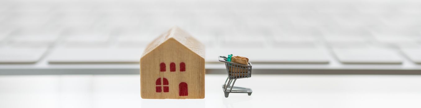 Online, E-Commerce and Technology Cover concept.  Close up of miniature shopping cart / trolley with wooden mini house toy and computer keyboard on white background.