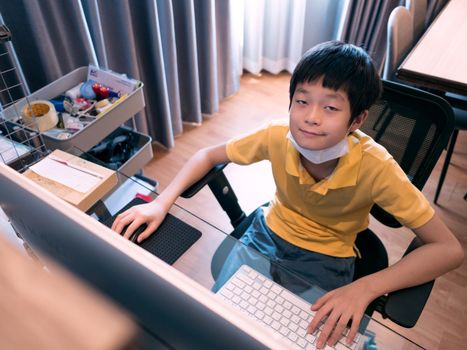cute asian boy hanging face mask on neck, looking at camera, sitting at desk in living room, using pc for studying online with monitor on blurred foreground, high angle view . Study online, stay home concept.