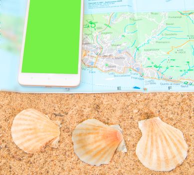 Green screen chromakey smartphone on the tourist map with shells on the sand, the phone screen can be replaced, the concept of tourism, top view.