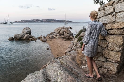 Beautiful woman in luxury summer dress enjoying peaceful seascape of Porto Rafael bay at Mediterranean sea of Costa Smeralda, Sardinia, Italy at dusk.