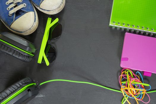 Back to school background with sneakers, headphones, sunglasses, colorful books and rubber band on black background. Flat lay, view from above