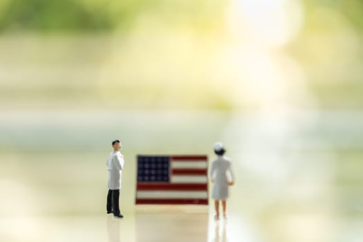 Global Healthcare, Coronavirus, Covid-19 Protection Concept. Doctor and nurse miniature figure people wearing face mask standing with United States of America (USA) flag pin and green nature background and copy space.