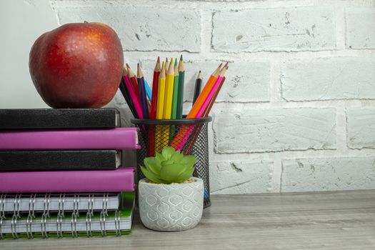Back to school concept with pile of books, red apple, potted plant and pencils against a white brick wall with free copy space