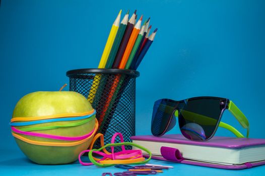 Back to school concept with book, green apple, rubber bands, sunglasses and pencils against a blue background with free copy space