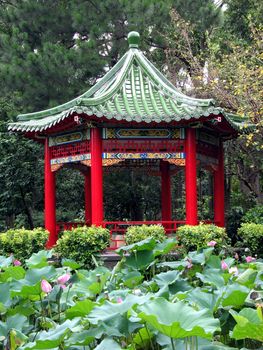 A Chinese pavilion with red columns and green roof amidst a pond with water lilies
