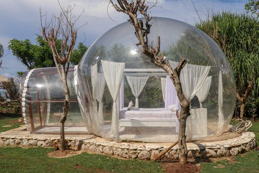 Bubble house with transparent walls. White wooden double bed with mosquito net inside bubble. Honeymoon in inflatable tent for tourists. Bubble hotel in tropical paradise on Bali island in Indonesia.
