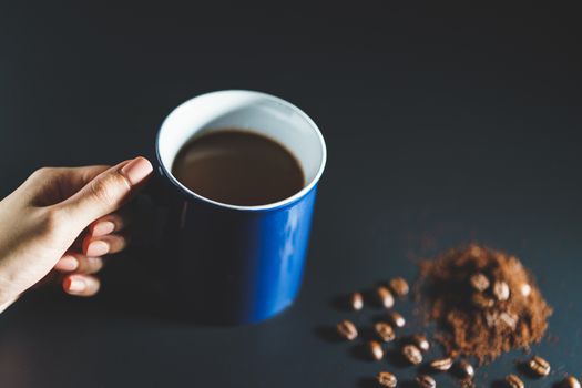 Hand holding a cup of coffee with coffee beans and powder on black background.