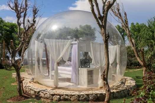 Romantic bubble hotel with transparent walls. White wooden double bed with mosquito net inside bubble house. Honeymoon in an inflatable tent. Tourist attraction in tropical paradise on Bali, Indonesia