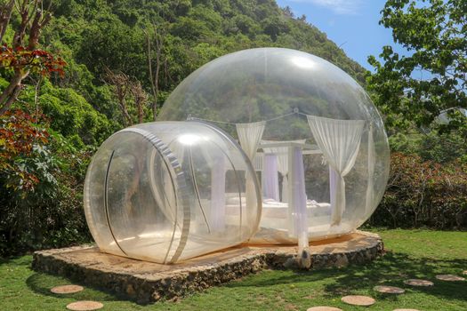 Glamping on Bali beach, in bubble house with transparent walls. White wooden double bed with mosquito net inside bubble. Honeymoon in an inflatable tent. Tourist attraction in tropical paradise.