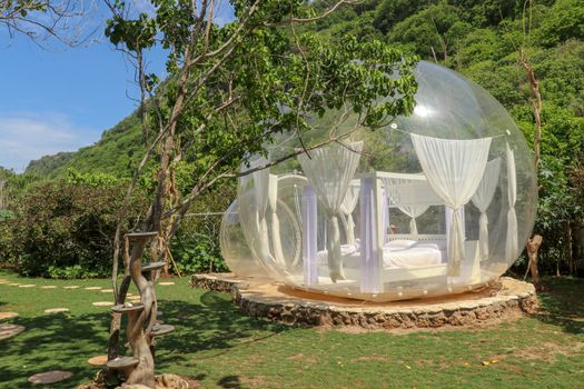 Bubble house with transparent walls. White wooden double bed with mosquito net inside bubble. Honeymoon in inflatable tent for tourists. Bubble hotel in tropical paradise on Bali island in Indonesia.