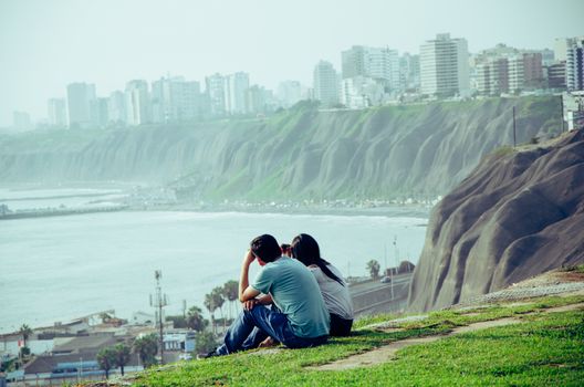 A couple sitting on the edge of the cliff on the coast of Lima - Peru facing the sea