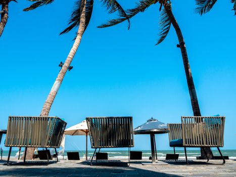 Modern chairs with round side table on balcony with coconut palm tree, beach umbrella and sea view near the beach on blue sky background.