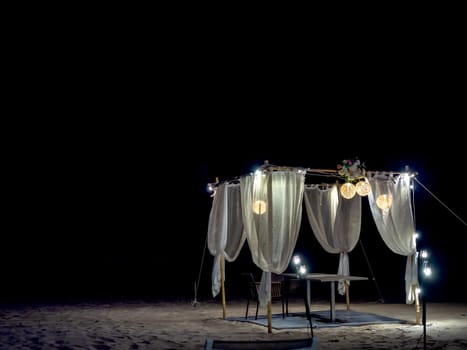 Romantic dining table, ornament with wood, lamp and white fabric on the beach at night on dark background.