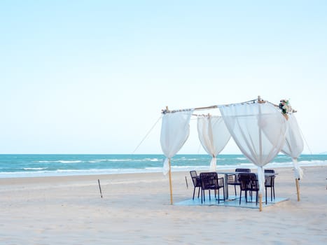 Romantic dining table, ornament with wood and white fabric on the beach on soft blue sky background.