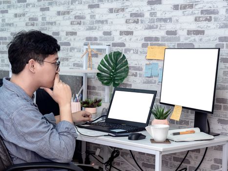 Asian man thinking and working with laptop computer with white blank screen and watching another monitor in his room, condominium. Work at home and business online concept.