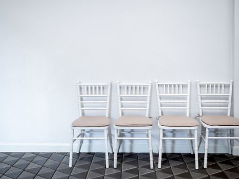 Row of clean white vintage wooden chairs with pads on modern stone tiles floor on white wall background with copy space.
