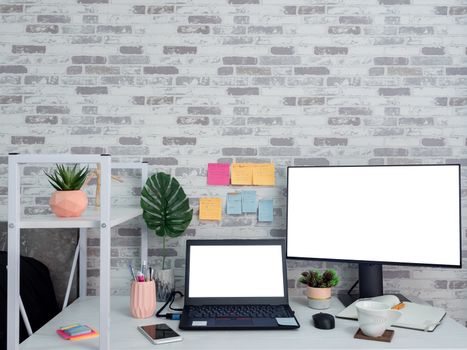 Work space in the room, White blank screen on laptop computer and monitor on desk decorating with houseplants, smartphone, notebook, coffee cup and notepad on the wallpaper.