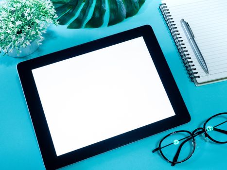 White blank space screen on tablet, pen, notebook, glasses and green leaves on blue background, top view.