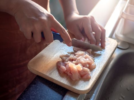 Close-up hand using a sharp knife, slice the chicken on a wooden cutting board near the window in room in condominium in the evening. Cooking at home.