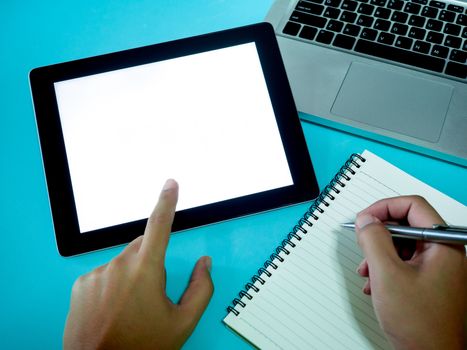 Hand pointing on white blank space screen on tablet and hand writing on notebook with laptop computer on blue background, top view.