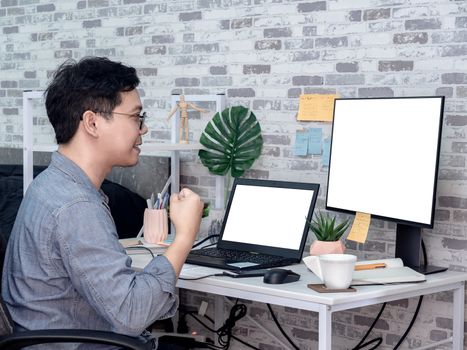 Asian man with success hand working with laptop computer with white blank screen and watching another monitor in his room, condominium. Work at home and business online concept.