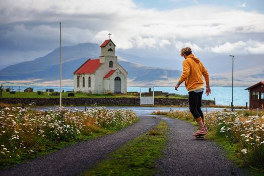 Young skater skateboarding towards a church and a cemetery in Iceland with a lake in the background.