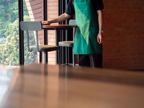 Waiter wearing green apron cleaning the round wooden table near the glass window and brick wall in modern loft style cafe with copy space.