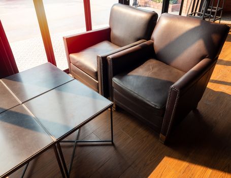 Brown leather sofa with wooden table on wooden floor near glass window in cafe on sunshine day.