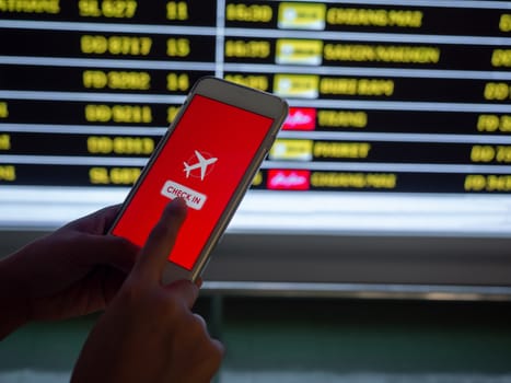 Flight check-in by mobile phone. Hand touching on smartphone screen to check-in for a flight in front of flight schedule board information background in airport.