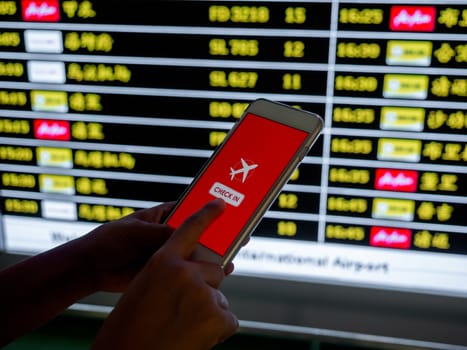 Flight check-in by mobile phone. Hand touching on smartphone screen to check-in for a flight in front of flight schedule board information background in airport.