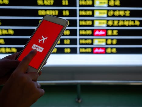 Flight check-in by mobile phone. Hand touching on smartphone screen to check-in for a flight in front of flight schedule board information background in airport.