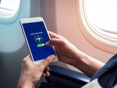 Flight mode concept. Hand holding white smartphone and turned on airplane mode on screen near the window on the airplane.