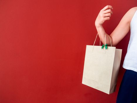 Woman's hand holding blank recycled paper bag decorate with little green bow on red wall background with copy space.