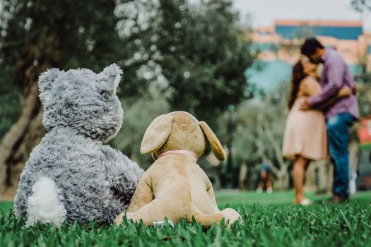 Two teddy bear toys sitting on the bench and hugging each other with nature background in the back, in the background a pregnant woman and her boyfriend kissing. Love song. Greeting or gift card design idea.