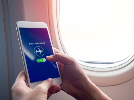 Flight mode concept. Hand holding white smartphone and turned on airplane mode on screen near the window on the airplane with copy space.