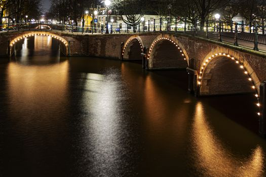 Illuminated Amsterdam canal bridge reflected on the calm canal water in Amsterdam at night