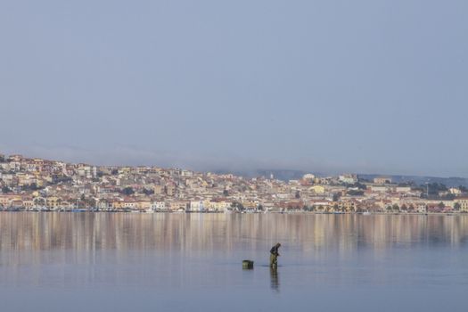 A fisherman at work bending in the flat sea water that reflects the houses of Sant'Antioco in Sardinia