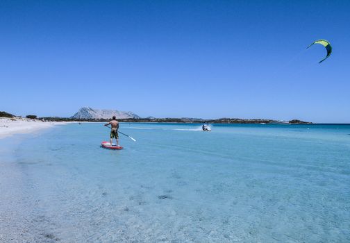 San Teodoro - La Cinta Beach - 2017 July - A young athlete paddle on stand up paddle on the crystalline sea of Sardinia, while a kitesurfer slides on the water beside him