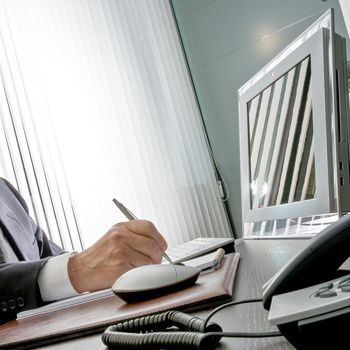 Manager at work. The expert hand of a businessman sitting at his desk, he holds the pen in front of his computer monitor that reflects the window vertical blinds of the office