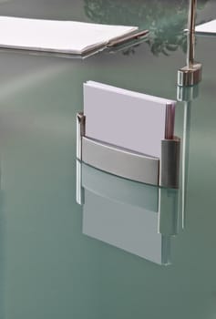 Anonymous business set. The elegant metal business card holder is reflected on the crystal of the desk in front of the notepad and the precious pen