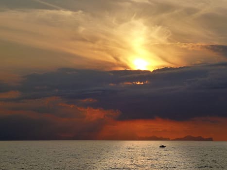 Spectacular sunset with red sky and clouds over the sea of Menorca in Spain with the silhouette of a boat on the bright reflection of the sun on the water