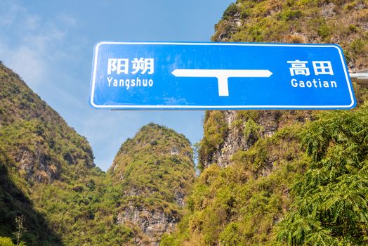Blue road sign with Yangshuo and Gaotian written in both chinese and romanian characters against karst hills and vegetation on a sunny day in Yangshuo, Guangxi province, China