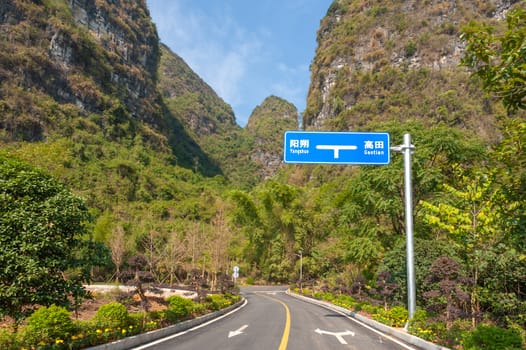 Empty road against karst hills and vegetation on a sunny day in Yangshuo with blue road sign with Yangshuo and Gaotian written in both chinese and romanian characters...