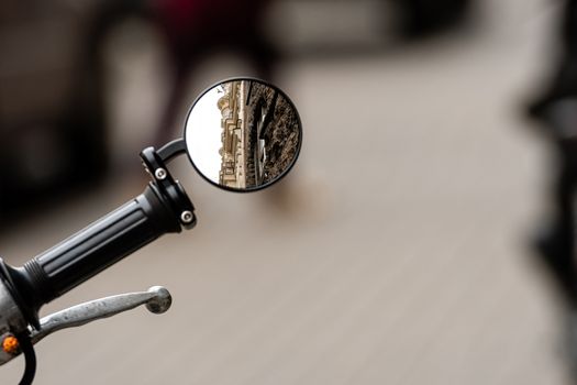 reflection of a ancient residental building in the rear view mirror of a motorcycle in front of a defocused sidewalk