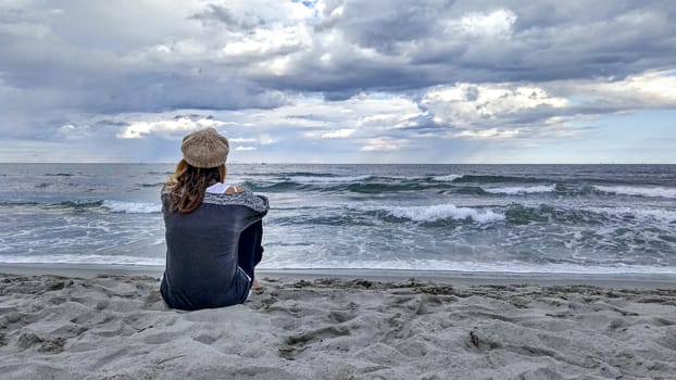 Young woman sitting by the sea with stormy sky, looks at the horizon thoughtfully