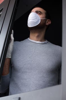 Man with face mask and gloves looking out the window. Stay at home concept.